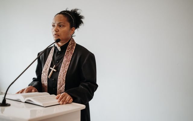 A female pastor delivers a sermon at a church podium with a Bible and microphone.