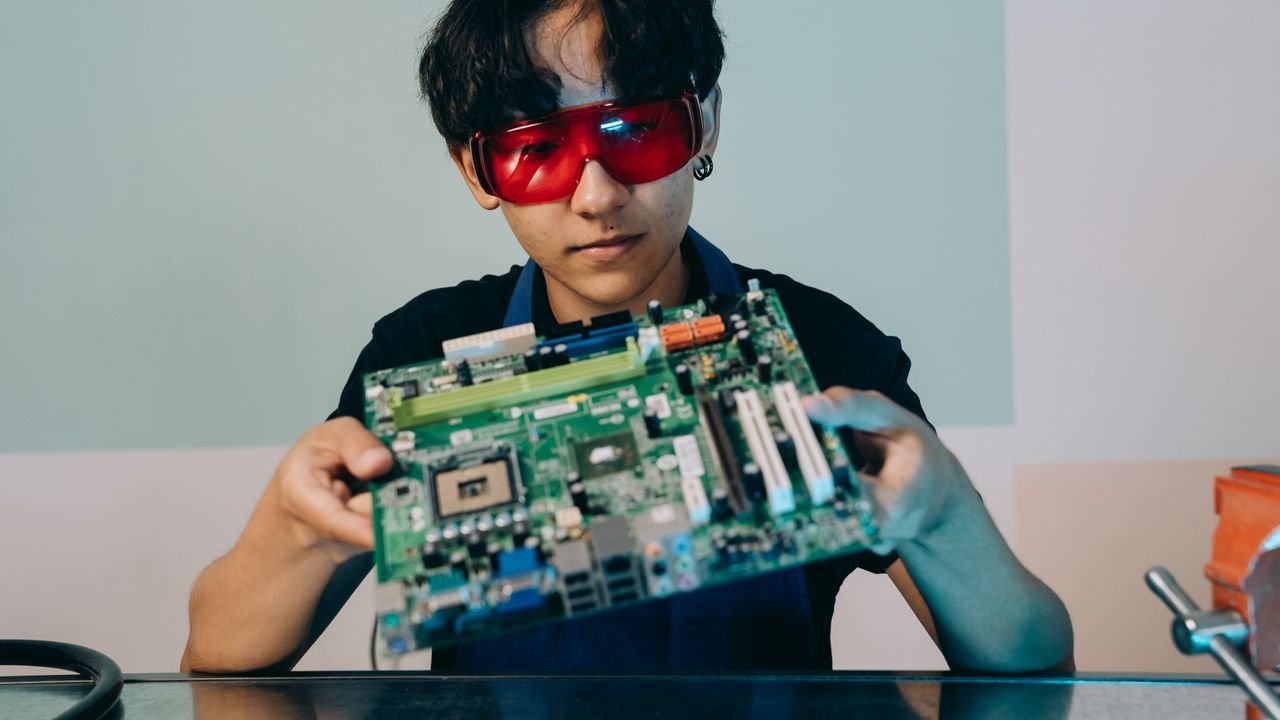 A focused young engineer holding a motherboard, wearing eye protection indoors.